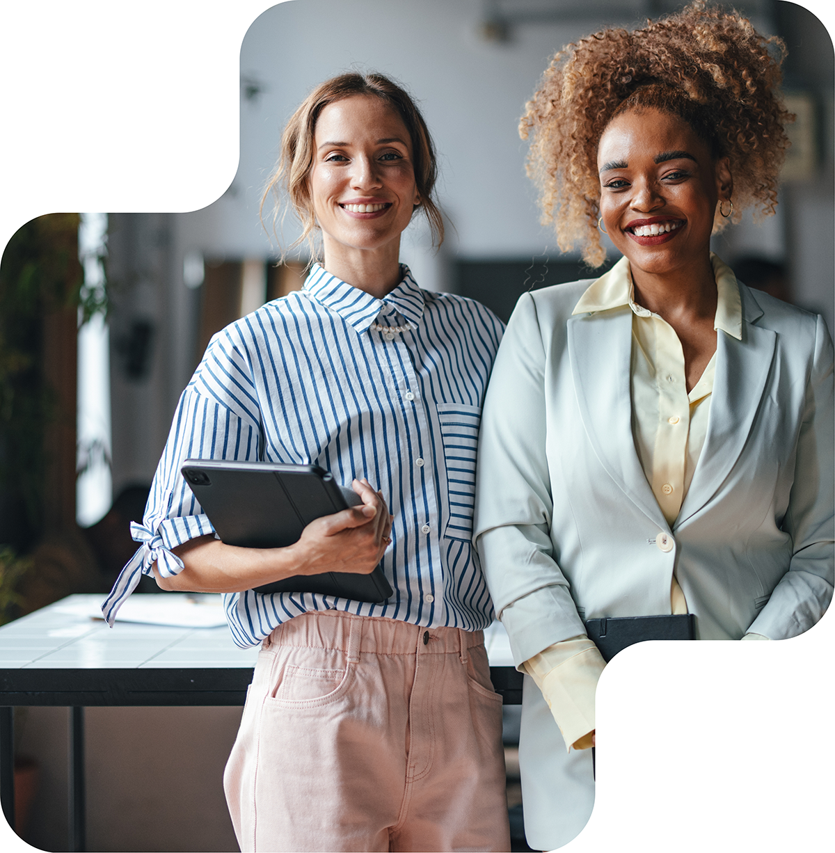 Two professional women smiling confidently, with one holding a tablet and the other a notebook, showcasing empowerment and collaboration in the workplace.