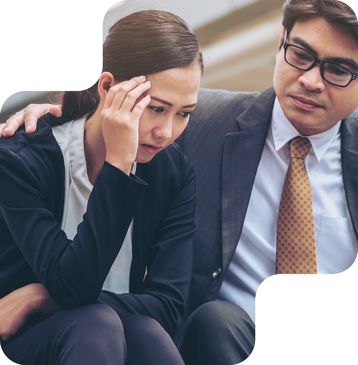 Image shows a worried woman sitting with her head in her hand, supported by a man in a suit and tie who has his arm around her. The man appears to be offering comfort or support to the woman.