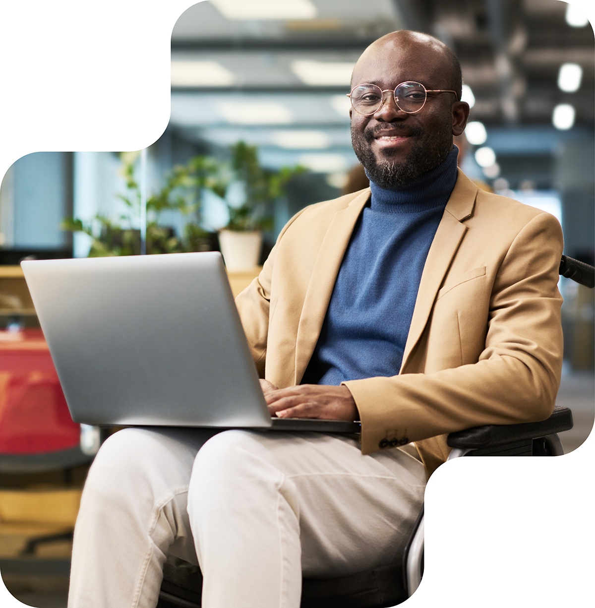 Image shows a man with glasses and a beard, smiling as he works on a laptop. He is wearing a suit jacket over a turtleneck sweater and is seated in a wheelchair. The man appears confident and engaged in his work.