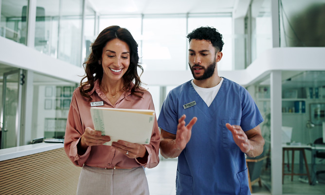 Two diverse healthcare professionals having an engaged conversation in a modern office setting, with a woman in business attire holding a tablet while a man in medical scrubs gestures expressively, demonstrating inclusive workplace communication