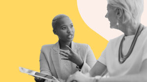 Image shows two women engaged in a conversation. One woman, with short cropped hair, is speaking animatedly while holding a tablet. The other woman, with short white hair, listens attentively. Both are dressed in professional attire. The background features a contrasting yellow and white color scheme. The image conveys a sense of engagement and professional interaction.