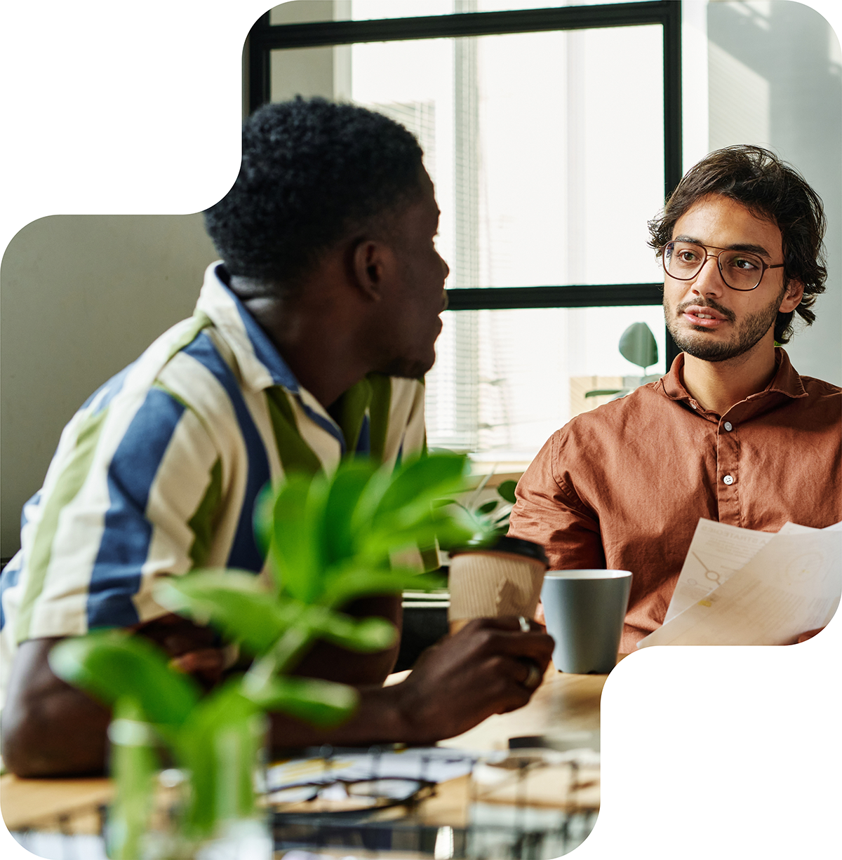 Two colleagues engaged in a serious conversation at a well-lit office table, surrounded by plants and documents, fostering an inclusive and collaborative work environment.