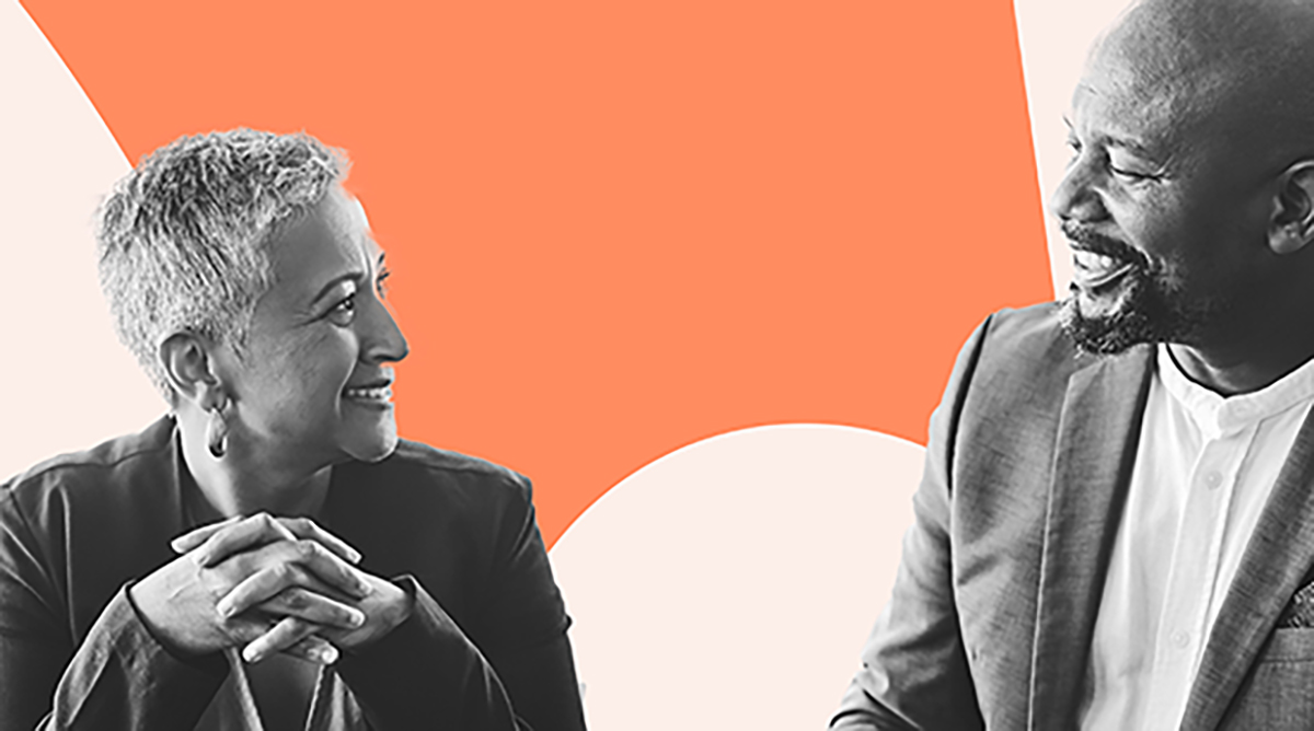 Two colleagues, a woman with short grey hair and a man with a beard, are engaged in a friendly and positive conversation against a vibrant orange and white background, highlighting a moment of connection and inclusivity in the workplace.