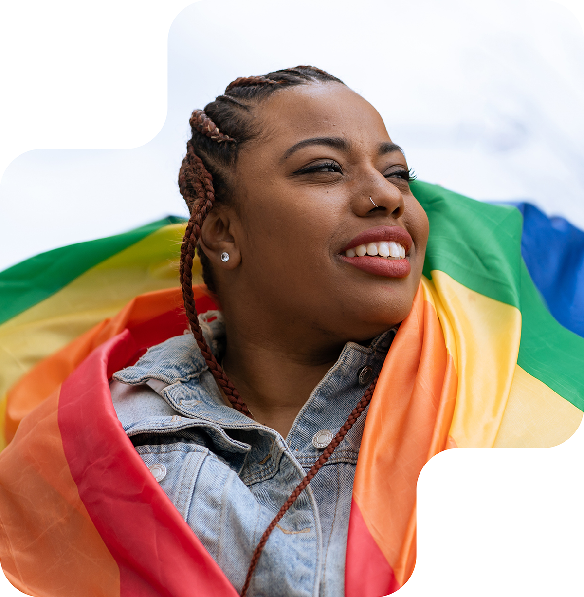 Image shows a young woman with braided hair, wearing a nose ring and earrings, smiling and looking into the distance. She appears content and optimistic. The background features a contrasting orange and beige color scheme. The woman is wrapped in a flag, adding a sense of pride and identity to the image.