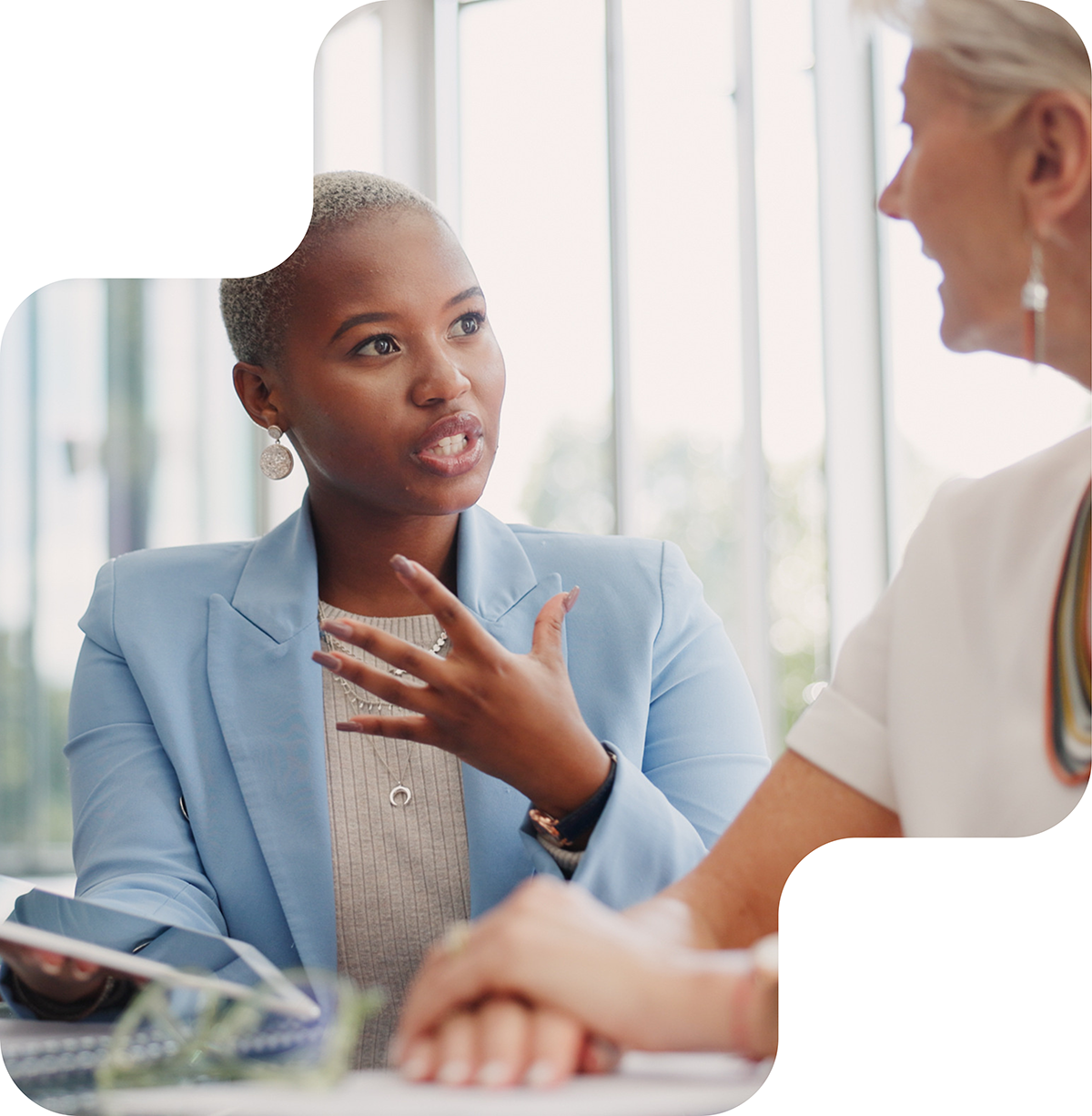 Image shows two women engaged in a conversation. One woman, with short cropped hair, is speaking animatedly while holding a tablet. The other woman, with short white hair, listens attentively. Both are dressed in professional attire. The background features a contrasting yellow and white color scheme. The image conveys a sense of engagement and professional interaction.