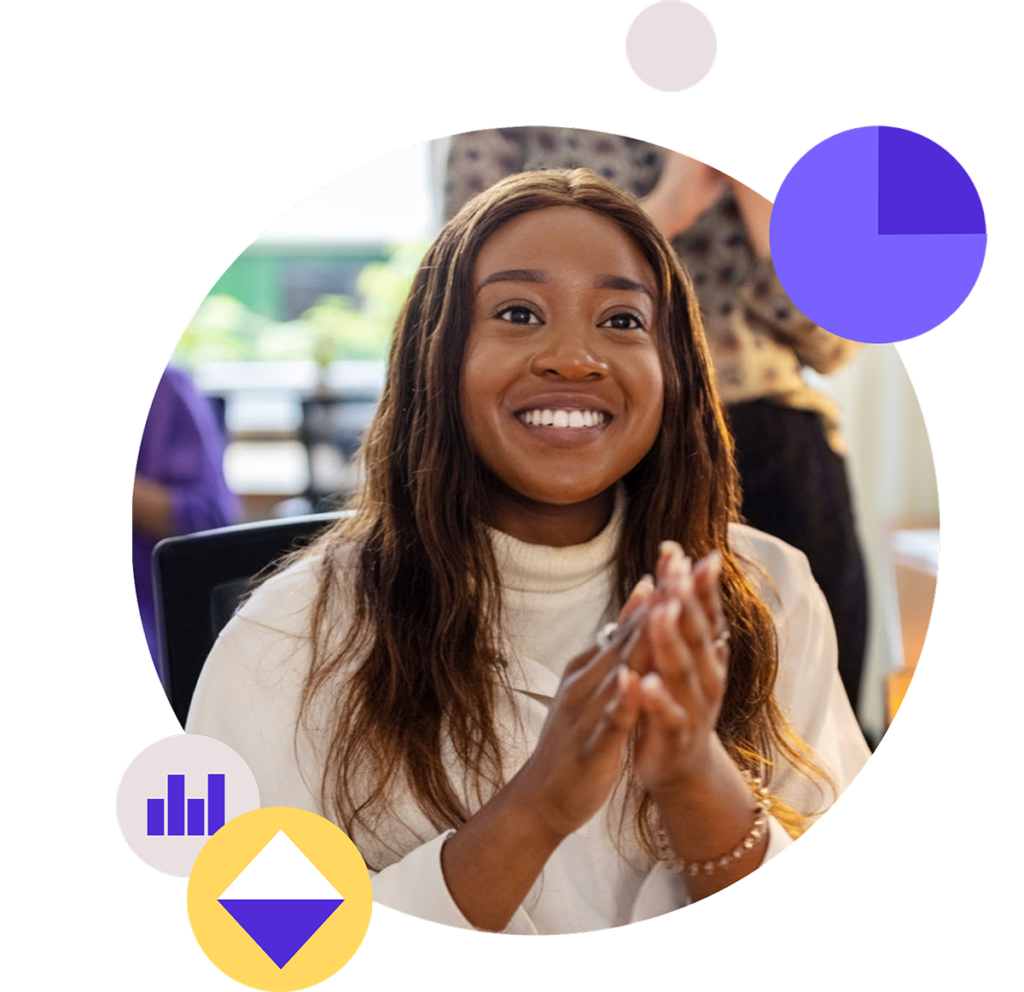 Smiling woman with long brown hair, wearing a white turtleneck top, sitting at a table and clapping her hands. She appears to be engaged and joyful, possibly in a team meeting or celebratory moment. Background includes softly blurred figures in a warm, well-lit indoor setting.